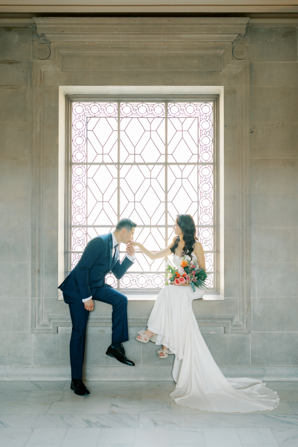 Bride and groom sit on window sill while the groom kisses the bride's hand during San Francisco city hall wedding bridals