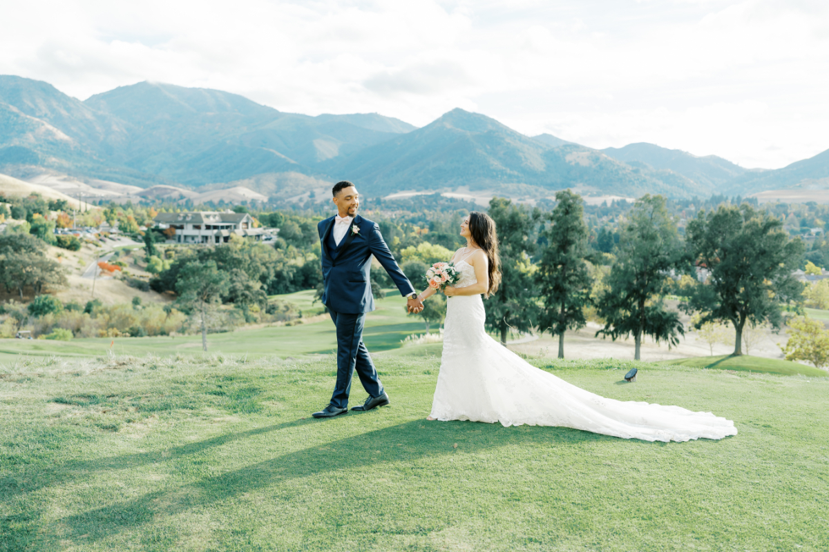 Bride and groom hold hands while walking along a hill with mountain outlines in the background