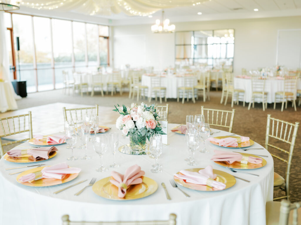Tables with white cloths, gold plates, and pink napkins with a floral arrangement in the middle. Reception hall at San Francisco wedding venues