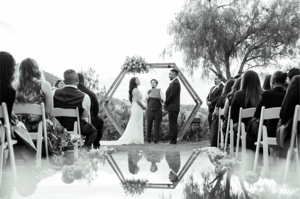 bride groom and officiant stand together underneath a wedding arch and are reflected in a puddle on the ground, black and white photo