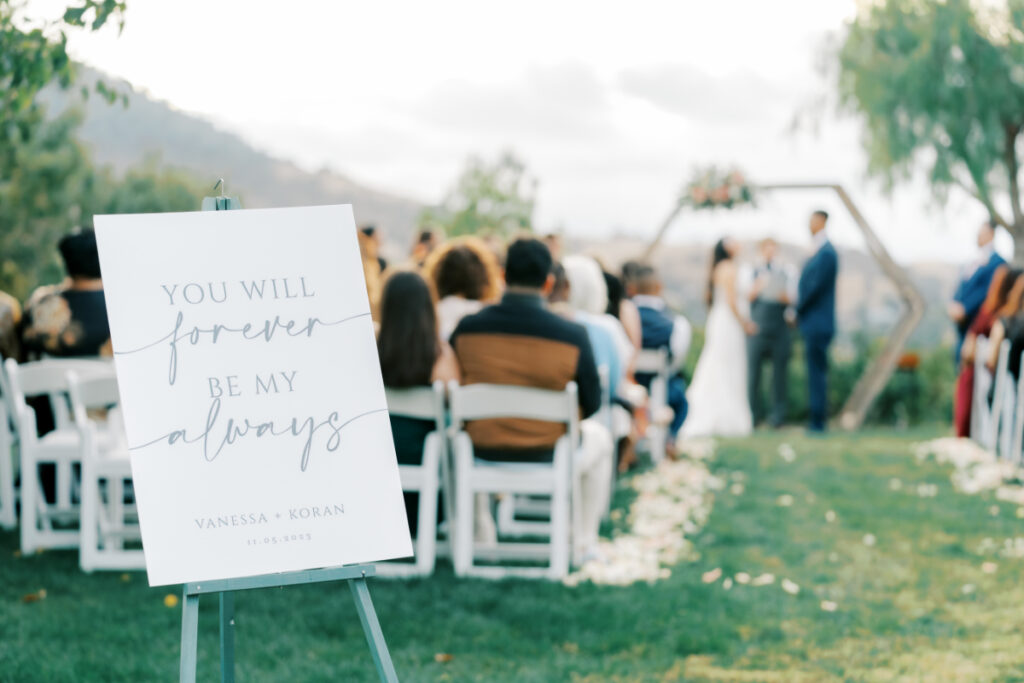 Sign says "you'll forever be my always" with a blurred background of guests watching the ceremony