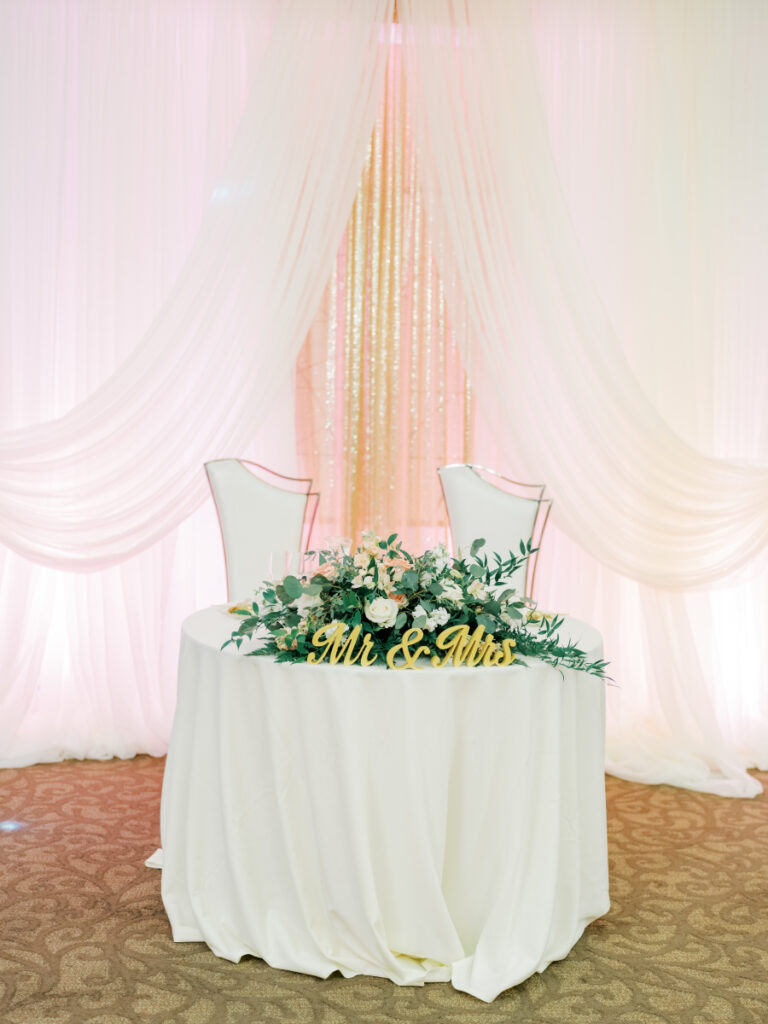 Table with white cloth and flowers with lots of greenery and signs that say "Mr & Mrs" with two white chairs and a pink curtain behind it. Bride and groom table at San Francisco wedding venue. 