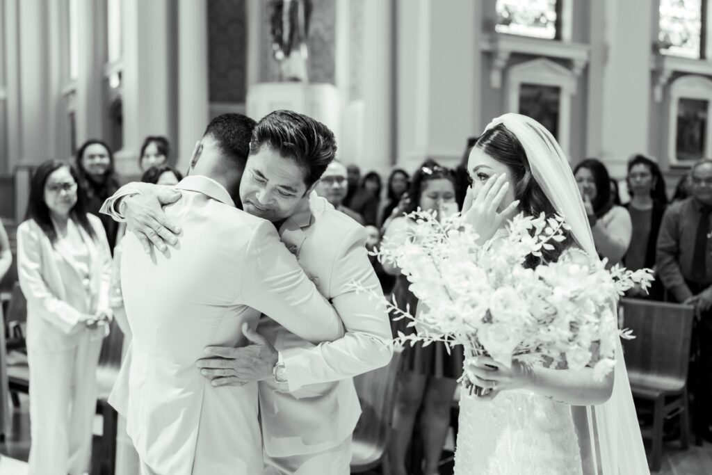Bride's father embraces his new son in law at the end of walking the bride down the isle and hands the bride over to the groom. The bride is seen wiping tears. Photo taken at Cathedral Basilica of St. Joseph
